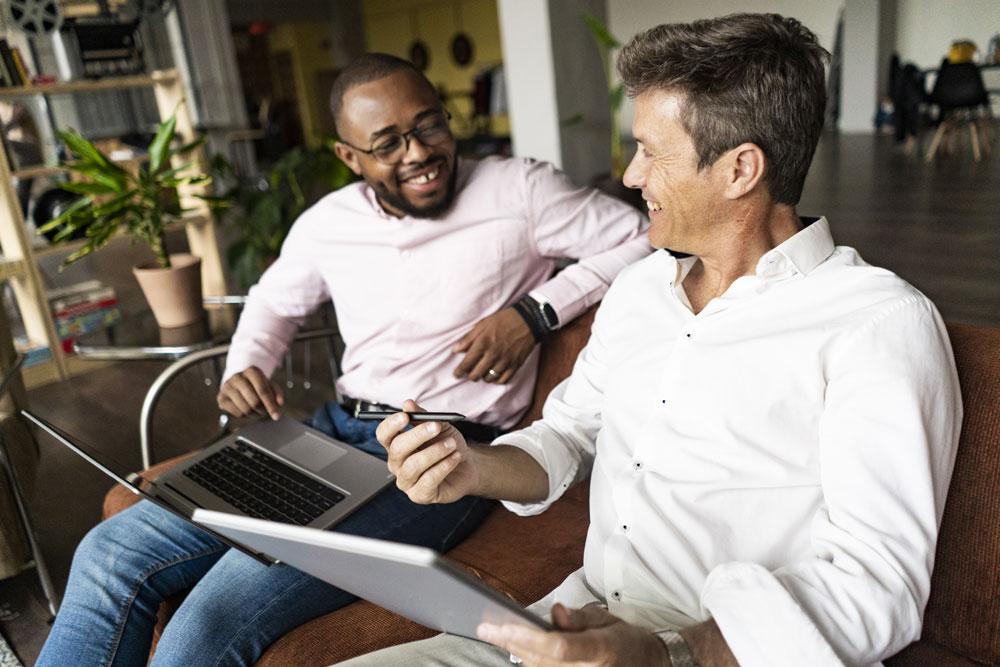 Two colleagues collaborating over a laptop and tablet in a relaxed workplace conversation